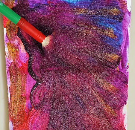 A colourful photo showing a snapshot of the process of a painting that is being created in glitter paint also showing the paintbrush being used.
